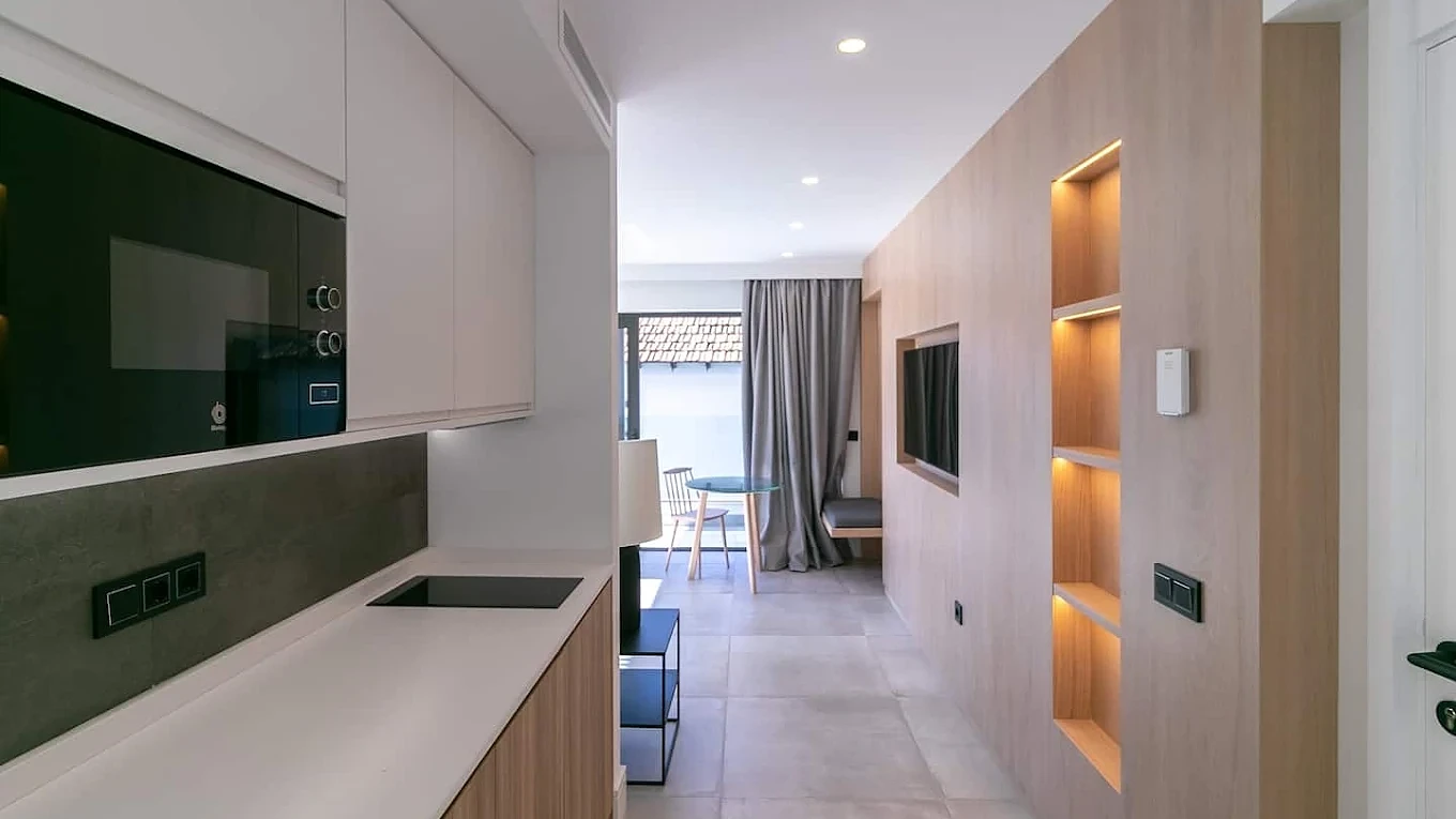 La misma cocina abierta al salón ya reformada, con muebles blancos, pared de roble con hornacinas iluminadas y suelo porcelánico